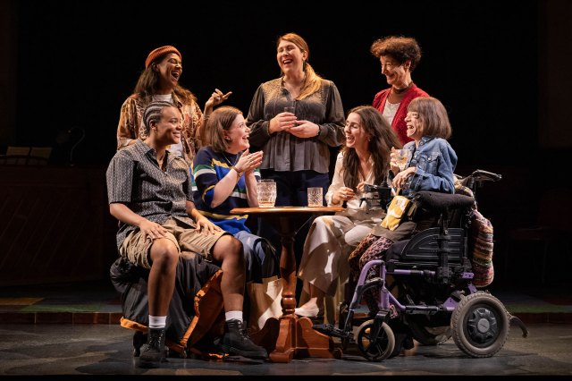 A group of actresses gathered around a pub table on stage