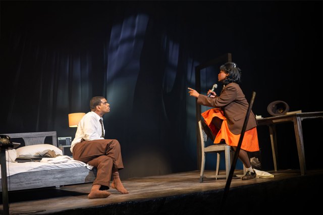 An actor in a white shirt and brown trousers and an actress in an orange maid's uniform in a discussion on a hotel room set on stage