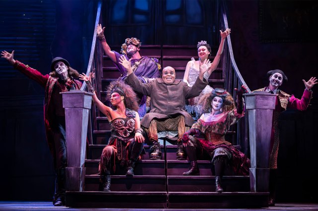 An actor sat on an onstage staircase, dressed as Uncle Fester, surrounded by cast members in gothic costumes