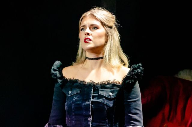 Erin Caldwell in Saving Mozart, she wears a black off shoulder ruffled top and choker, looking into the distance contemplatively