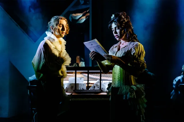 An actor and an actress holding sheet music, dressed in stylised period costumes on stage