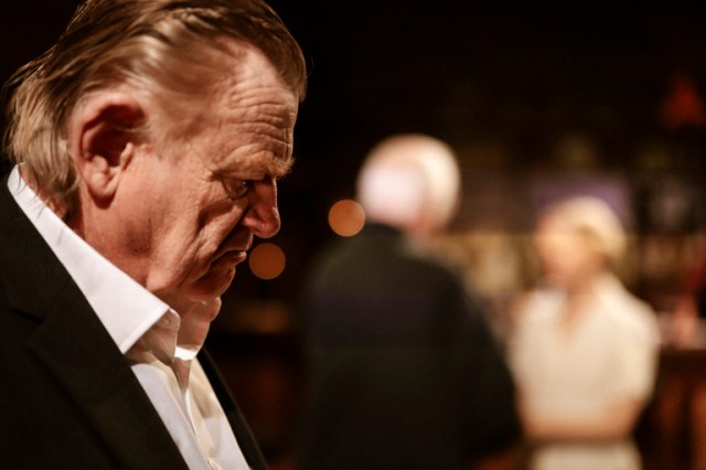 Brendan Gleeson's side profile as the actor looks down, his face looks gloomy and out of focus behind him are blurred figures in conversation. It looks dark, in a pub setting.