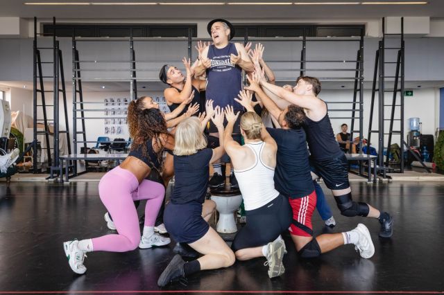 The cast of The Producers in rehearsals The cast of The Producers in rehearsals, a man stands tall centrally as performers kneel around him reaching up lovingly