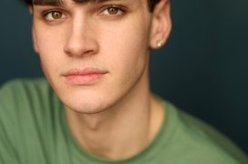 Close-up portrait of a young man with dark wavy hair and brown eyes, wearing a green shirt against a dark teal background, looking at the camera.