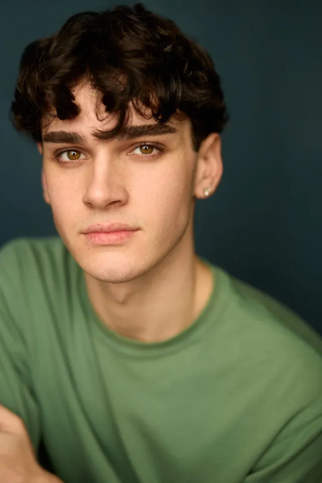 Close-up portrait of a young man with dark wavy hair and brown eyes, wearing a green shirt against a dark teal background, looking at the camera.