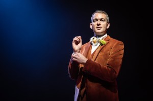 Stage performer in a brown velvet suit with a boutonniere, under blue spotlight, adjusting a cufflink before a show.