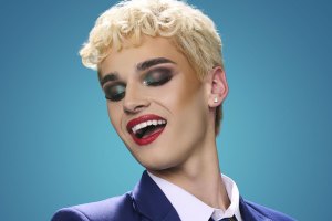 Portrait of a person with short platinum blonde hair, bold makeup, and bright red lipstick, wearing a blue blazer and white shirt against a teal gradient background, smiling with eyes closed.