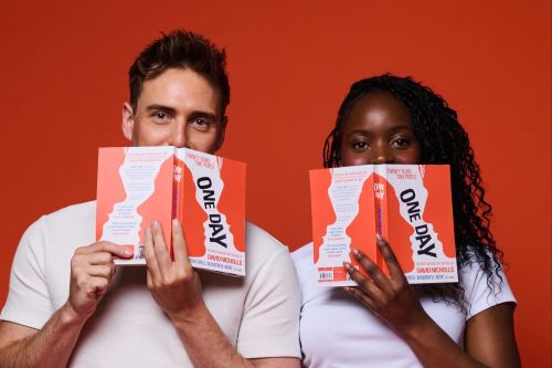Two people sit against an orange background, each holding a bright orange 'ONE DAY' book that covers part of their faces, wearing white tops.
