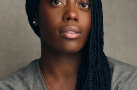 Close-up portrait of a woman with long black braids, wearing a gold pendant necklace and a gray top, neutral backdrop.