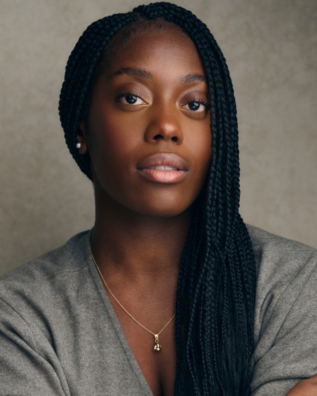 Close-up portrait of a woman with long black braids, wearing a gold pendant necklace and a gray top, neutral backdrop.