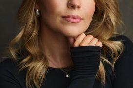 Close-up portrait of a blonde woman with green eyes and a nose stud, resting her chin on her hand in a black top.