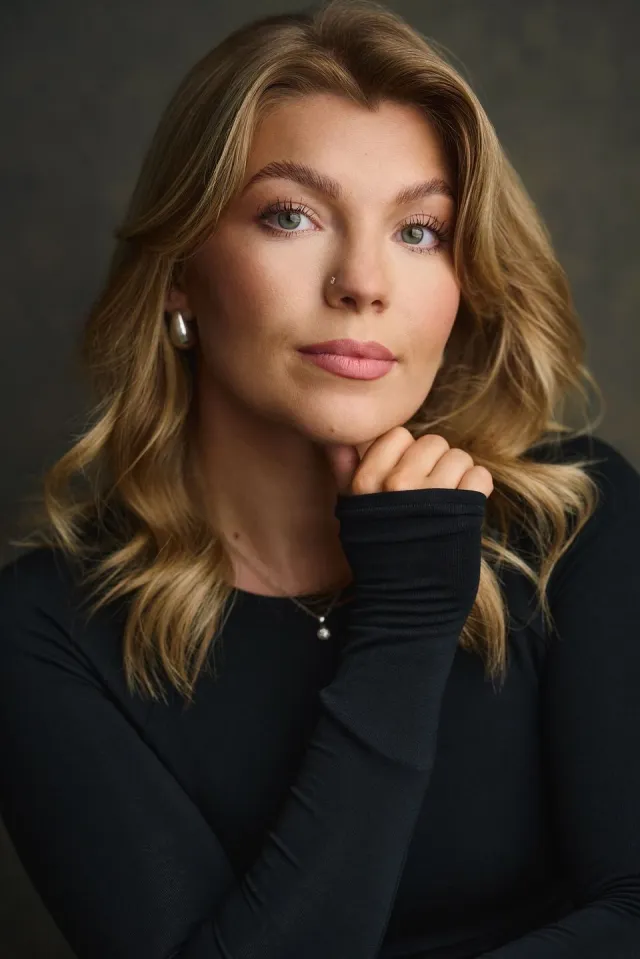 Close-up portrait of a blonde woman with green eyes and a nose stud, resting her chin on her hand in a black top.