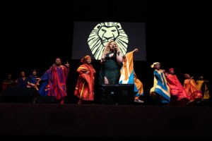 Lead female singer in a sparkling green dress performs on a dark stage, flanked by dancers in bright, striped robes with a lion emblem projected behind them.
