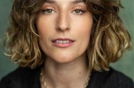 Portrait of a woman with shoulder-length wavy brown hair, green eyes, wearing a black blouse and a gold chain necklace.