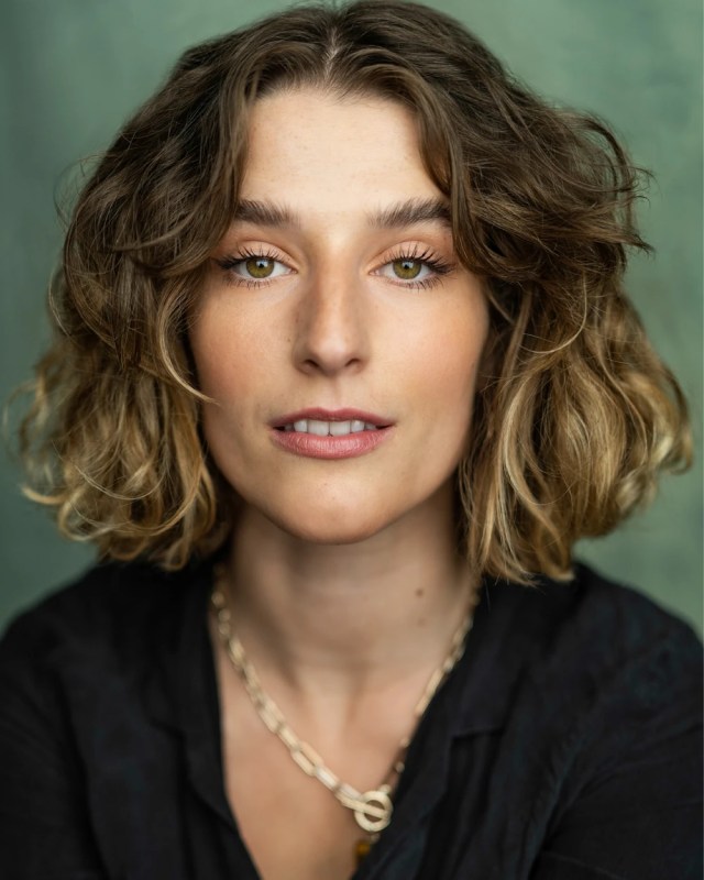 Portrait of a woman with shoulder-length wavy brown hair, green eyes, wearing a black blouse and a gold chain necklace.