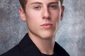 Male head-and-shoulders portrait of a young man with short curly brown hair wearing a black jacket against a textured gray backdrop, neutral expression—suitable for a professional headshot or profile photo.