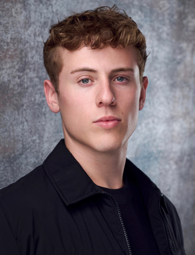 Male head-and-shoulders portrait of a young man with short curly brown hair wearing a black jacket against a textured gray backdrop, neutral expression—suitable for a professional headshot or profile photo.