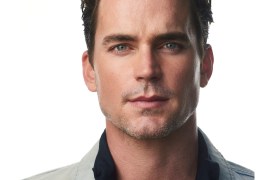 Close-up of a man with blue eyes and dark hair, wearing a light denim jacket and a dark shirt, neutral background.