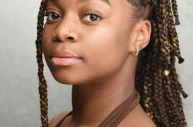 Portrait of a young woman with braided hair and a nose stud, wearing a brown halter top, looking toward the camera.
