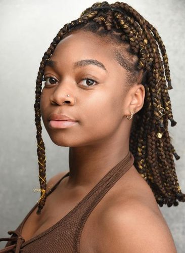 Portrait of a young woman with braided hair and a nose stud, wearing a brown halter top, looking toward the camera.