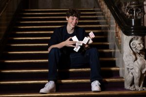 Young man sitting on a grand stairway, mid-card trick, smiling with cards in his hands.