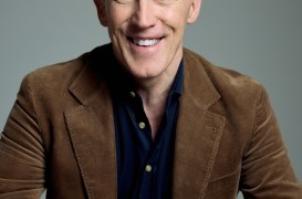 Smiling middle-aged man with gray hair wearing a brown corduroy jacket and dark shirt, seated at a table with hands resting on it.
