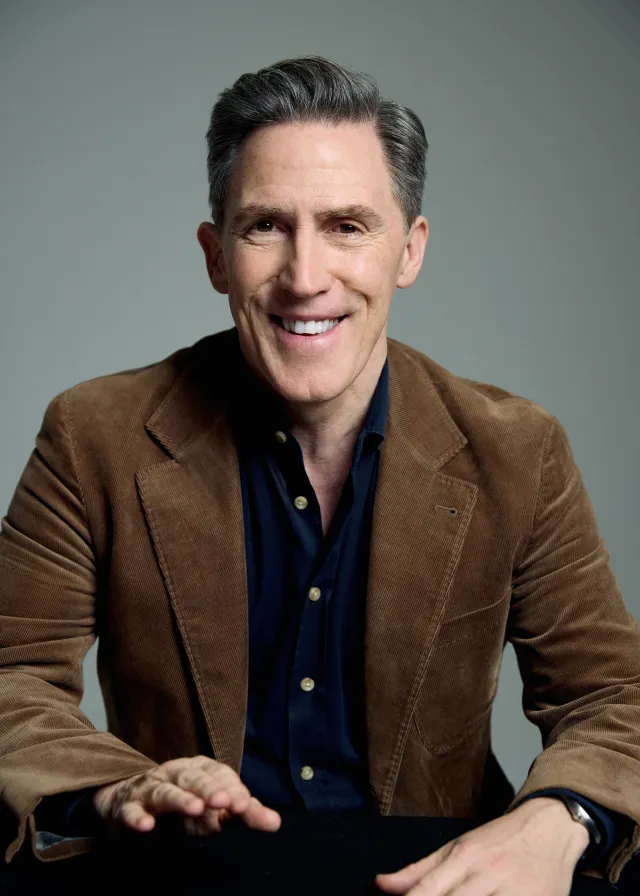 Smiling middle-aged man with gray hair wearing a brown corduroy jacket and dark shirt, seated at a table with hands resting on it.