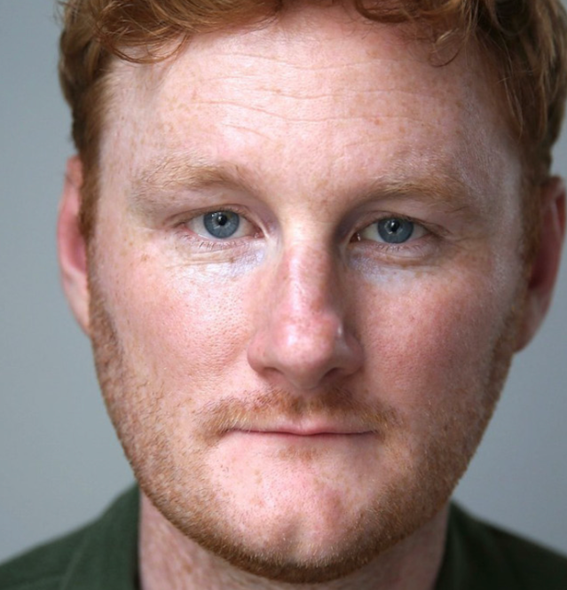 Close-up portrait of a man with red hair and blue eyes, neutral expression.