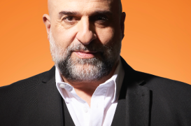 Bald man with a gray beard in a black suit and white shirt, against an orange background.