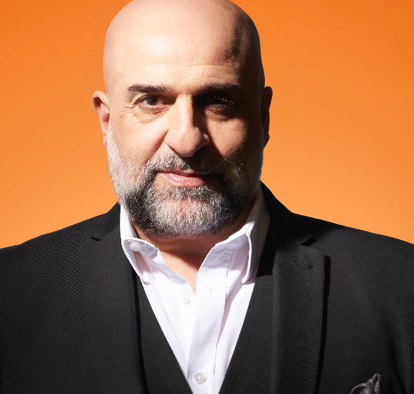 Bald man with a gray beard in a black suit and white shirt, against an orange background.
