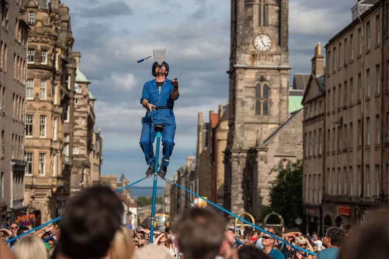 Street performer in blue overalls balances on a tall unicycle above a crowd on a historic city street, with a clock tower in the background.