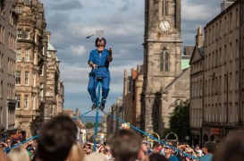 Street performer in blue overalls balances on a tall unicycle above a crowd on a historic city street, with a clock tower in the background.