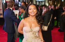 Smiling woman on a red carpet in a pale pink strapless gown with gold embroidery, posing for photos amid a crowd at night.