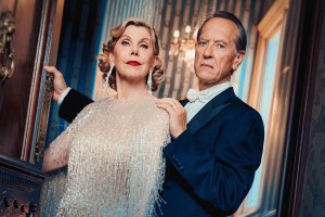 A stylish older couple posing at a grand doorway: the woman in a shimmering fringe gown with long earrings, the man in a dark tuxedo and white bow tie, both looking confident.