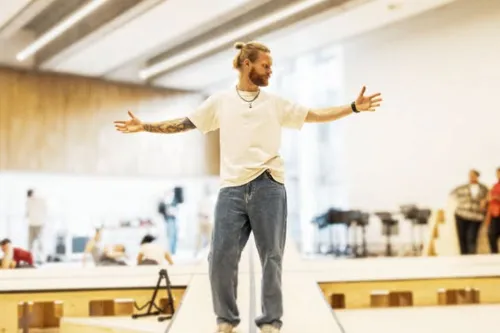 Man with arms outstretched stands on a small stage in a bright indoor space, seemingly speaking or presenting to others in the background.