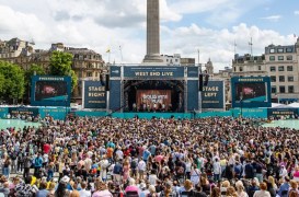 Large crowd gathers for a West End Live concert in Trafalgar Square, London, watching a big stage with banners and screens.