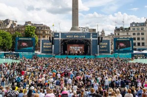 Large crowd gathers for a West End Live concert in Trafalgar Square, London, watching a big stage with banners and screens.